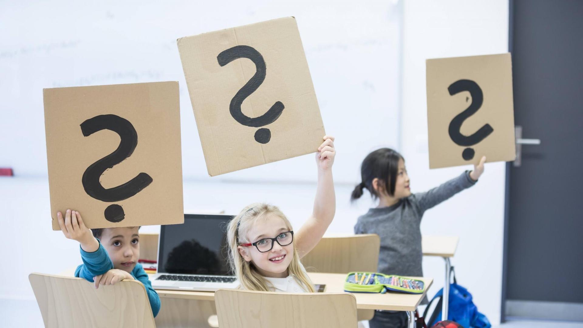 Grundschüler in einer Klasse mit Schildern, auf denen Fragezeichen gemalt sind. Grundschüler in einer Klasse mit Schildern, auf denen Fragezeichen gemalt sind.
