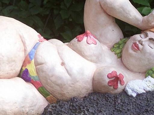 Skulptur einer Badenixe von der Künstlerin Leda Astorga, die eine füllige, liegende Frau beim Sonnenbaden zeigt, wobei über ihren Brustwarzen große, rote Blumen gelegt sind und sie eine bunte Badekappe trägt.