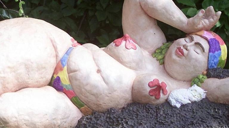 Skulptur einer Badenixe von der Künstlerin Leda Astorga, die eine füllige, liegende Frau beim Sonnenbaden zeigt, wobei über ihren Brustwarzen große, rote Blumen gelegt sind und sie eine bunte Badekappe trägt. Skulptur einer Badenixe von der Künstlerin Leda Astorga, die eine füllige, liegende Frau beim Sonnenbaden zeigt, wobei über ihren Brustwarzen große, rote Blumen gelegt sind und sie eine bunte Badekappe trägt.