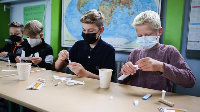 Kinder mit Gesichtsmasken in einem Klassenzimmer führen einen Coronatest durch.
