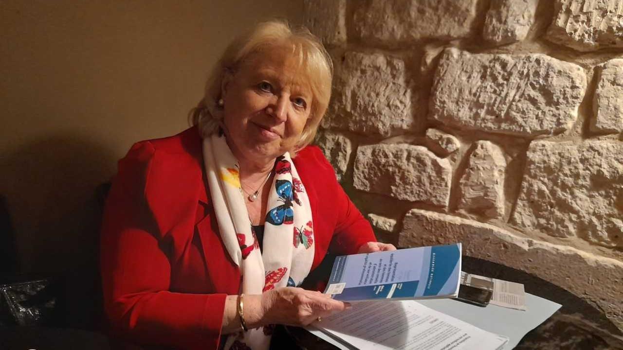 Eine blonde Frau mit rotem Blazer hat vor sich auf einem ein Broschüre und Papiere liegen. Eine blonde Frau mit rotem Blazer hat vor sich auf einem ein Broschüre und Papiere liegen.