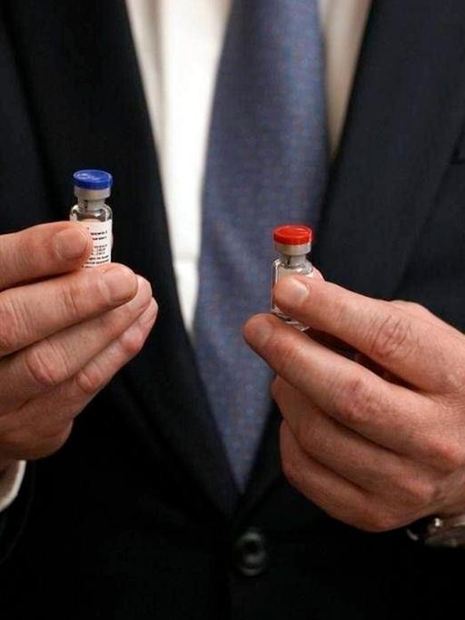 Nahaufmen eines Mannes im Anzug, der in seinen händen ein kleines Fläschchens hält, die Kappe des rechten ist blau, die des linken rot.