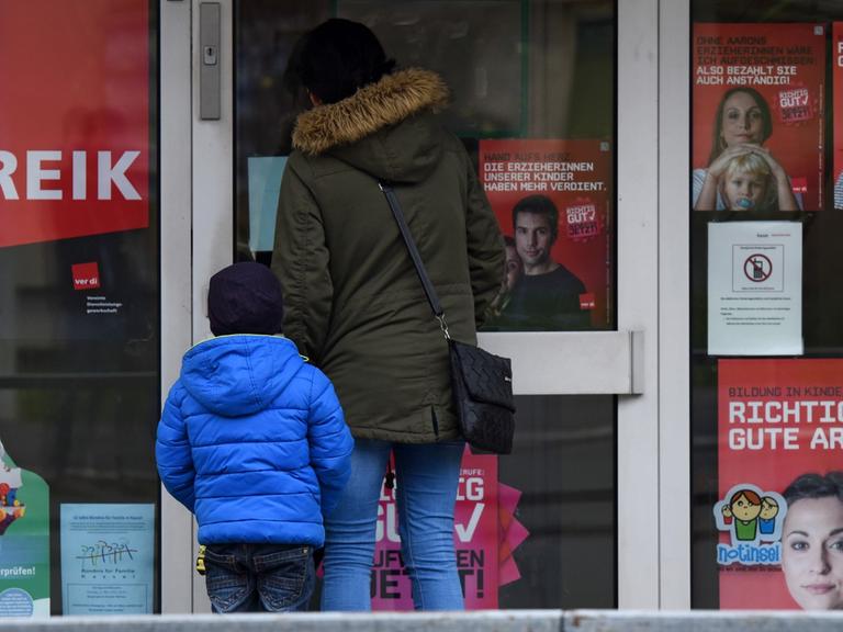 Eine Mutter steht mit ihrem Kind in Kassel (Hessen) vor einer geschlossenen Kita, an der Streik-Hinweise hängen