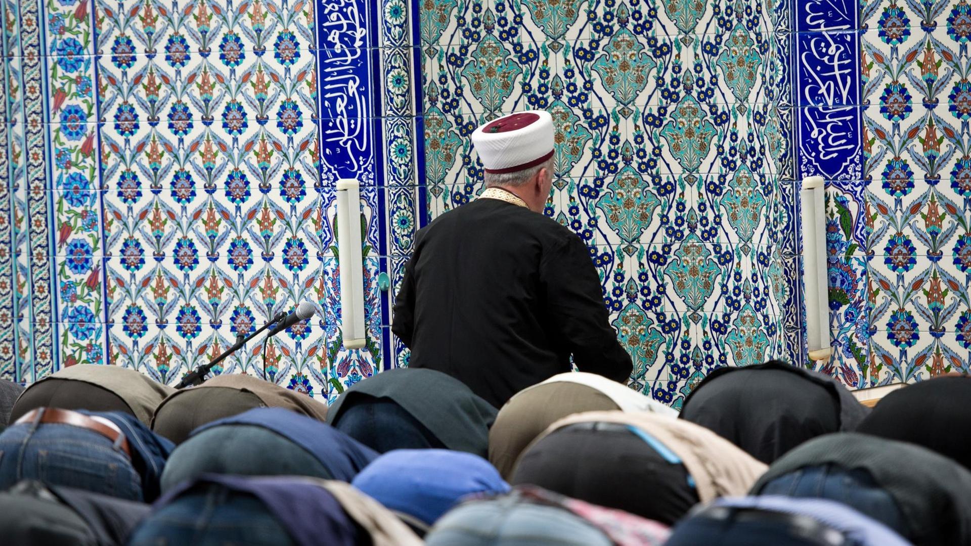 Viele Betende senken den Kopf auf den Boden. Vor ihnen sieht man von hinten den Imam vor einer Wand mit blauen Ornamenten. Viele Betende senken den Kopf auf den Boden. Vor ihnen sieht man von hinten den Imam vor einer Wand mit blauen Ornamenten.