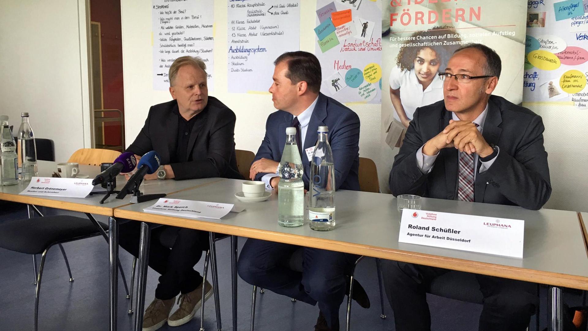 Drei Männer mittleren Alters sitzen an einem Tisch mit Mikrofonen vor sich. Sie werden gleich eine Pressekonferenz geben.