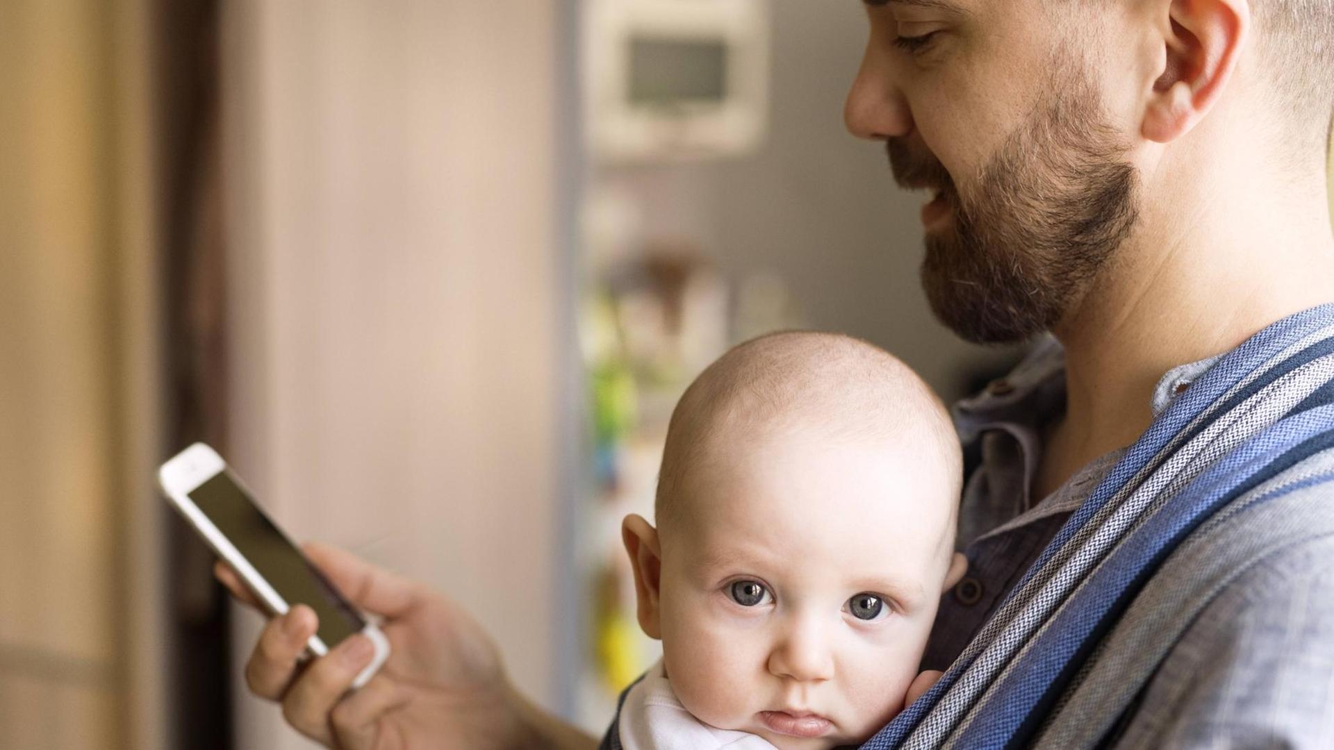 Ein Vater trägt ein Baby an der Brust und schaut dabei auf sein Smartphone Ein Vater trägt ein Baby an der Brust und schaut dabei auf sein Smartphone