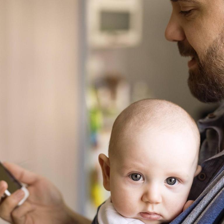 Ein Vater trägt ein Baby auf der Brust und schaut auf sein Smartphone