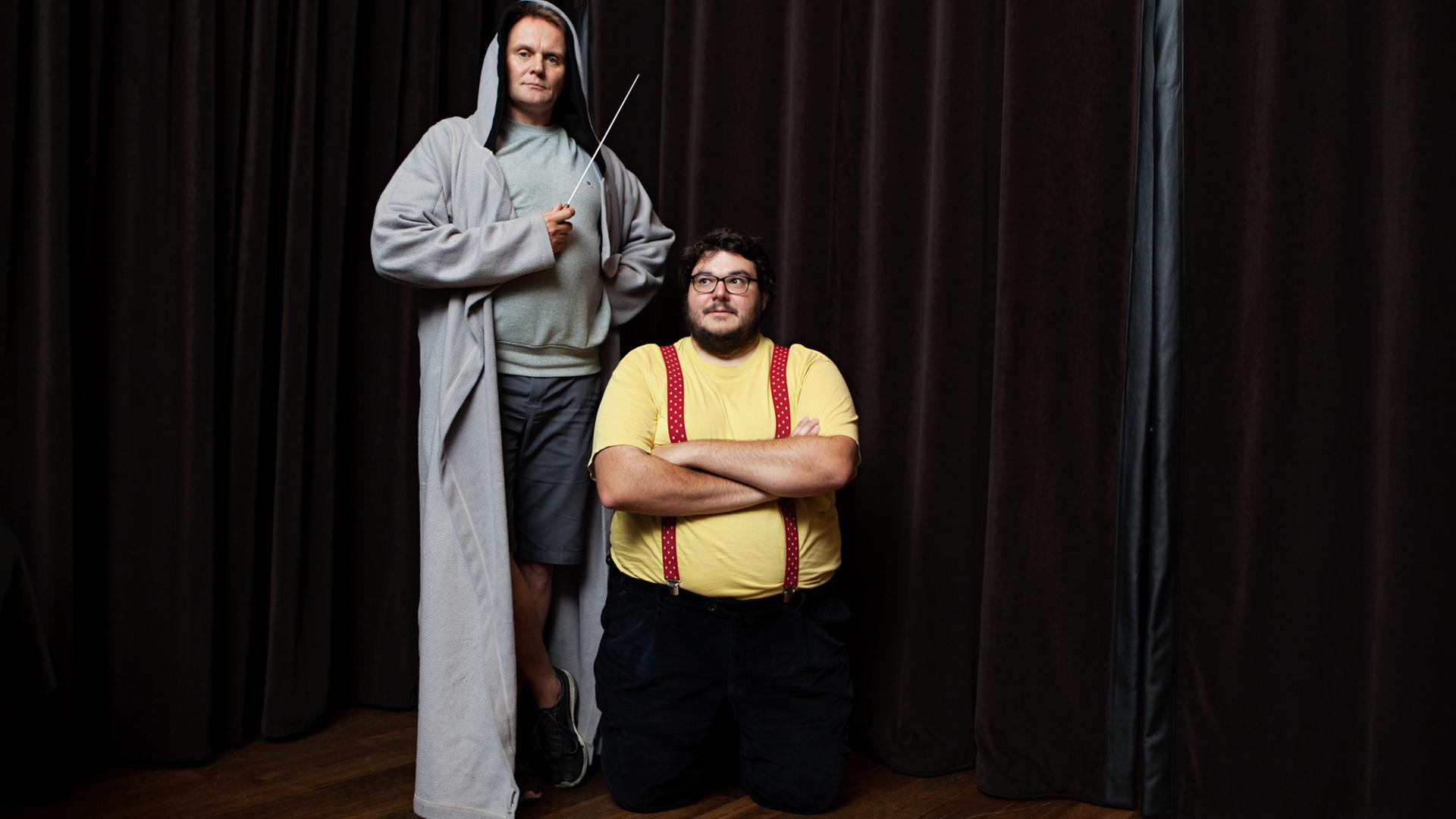 Das Bild zeigt Devid Striesow, der einen Kaputzenmantel trägt und auf einem Podest steht, einen Taktstock hält. Rechts neben ihm steht Axel Ranisch mit verschränkten Armen. Das Bild zeigt Devid Striesow, der einen Kaputzenmantel trägt und auf einem Podest steht, einen Taktstock hält. Rechts neben ihm steht Axel Ranisch mit verschränkten Armen.