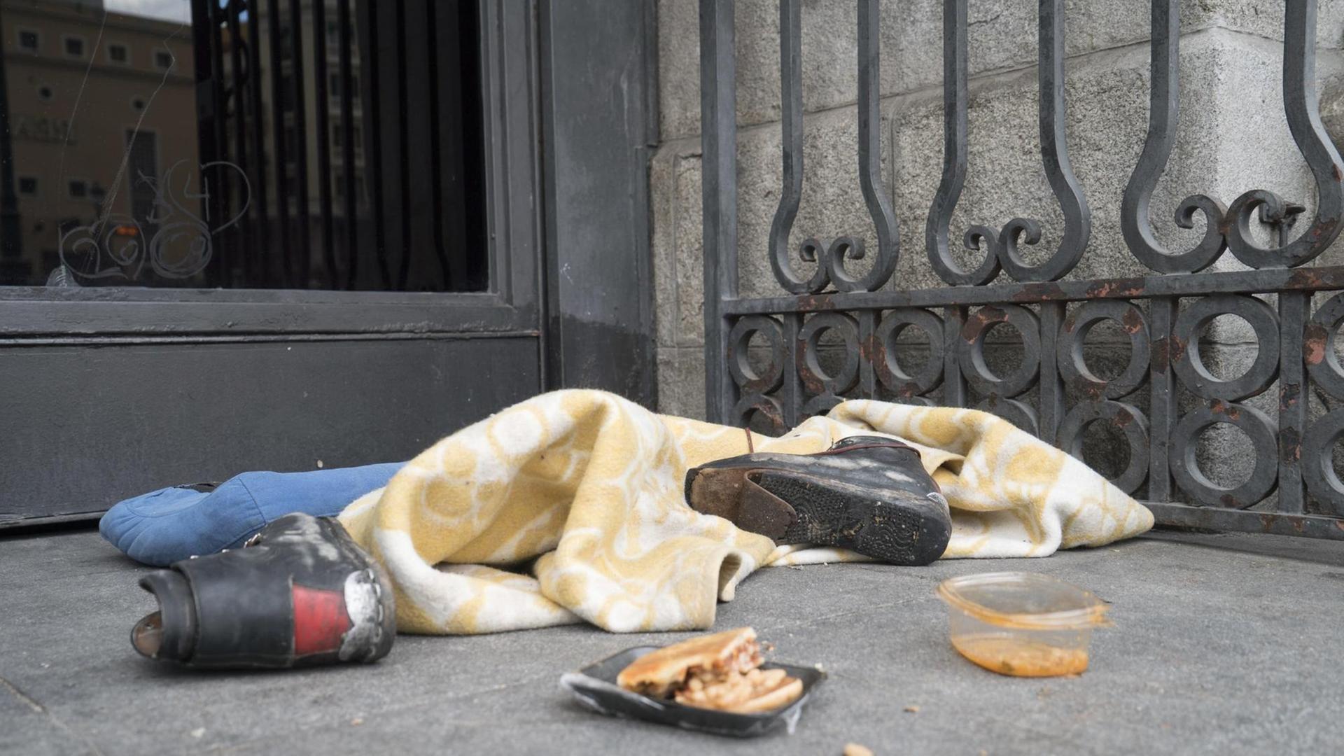 Lagerplatz eines Obdachlosen mit zerrissenen Schuhen, Essensresten und einer Decke vor einem Geschäft in Madrid. Lagerplatz eines Obdachlosen mit zerrissenen Schuhen, Essensresten und einer Decke vor einem Geschäft in Madrid.