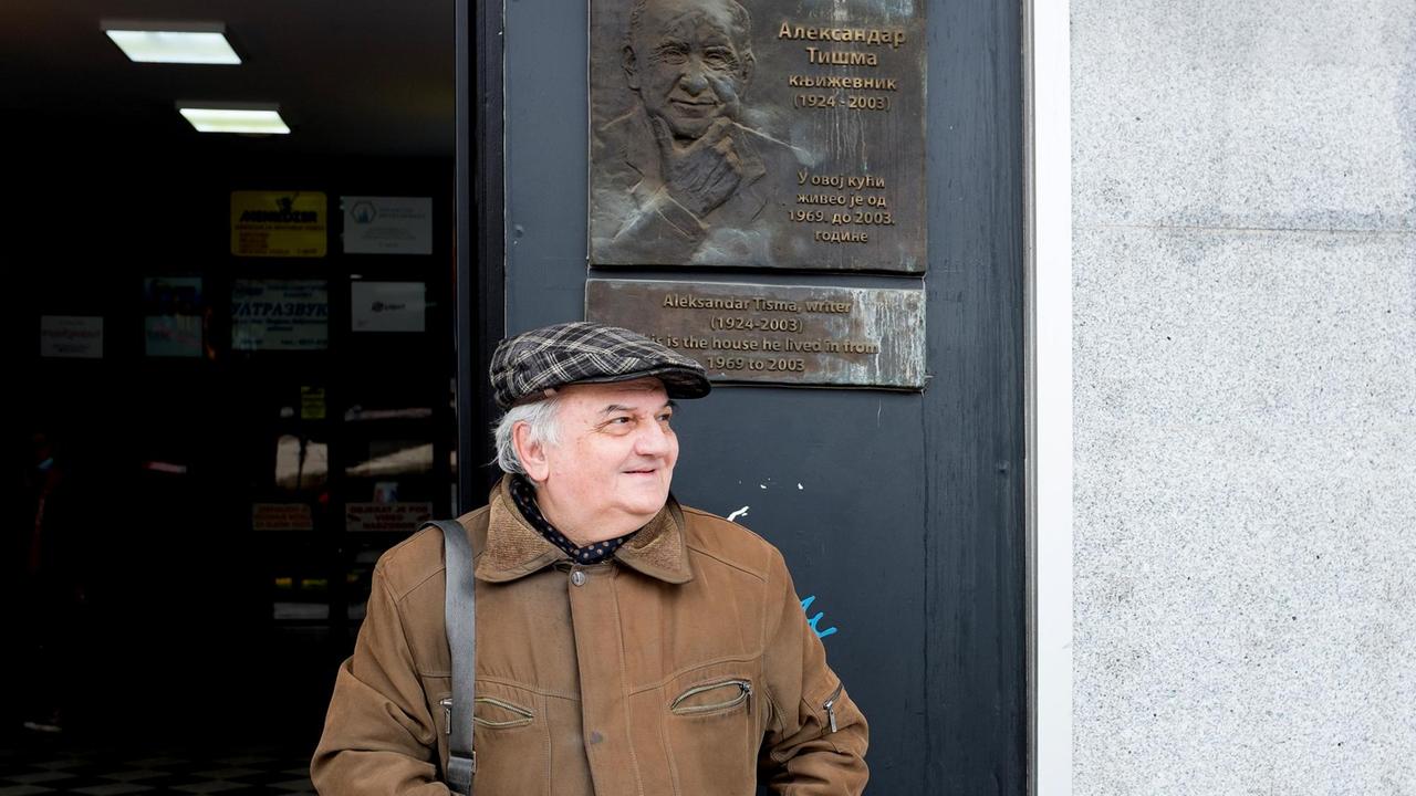 Andrej Tišma mit Schiebermütze steht lächelnd an einem Hauseingang, über ihm eine Hinweistafel zu seinen Vater Andrej Tišma mit Schiebermütze steht lächelnd an einem Hauseingang, über ihm eine Hinweistafel zu seinen Vater