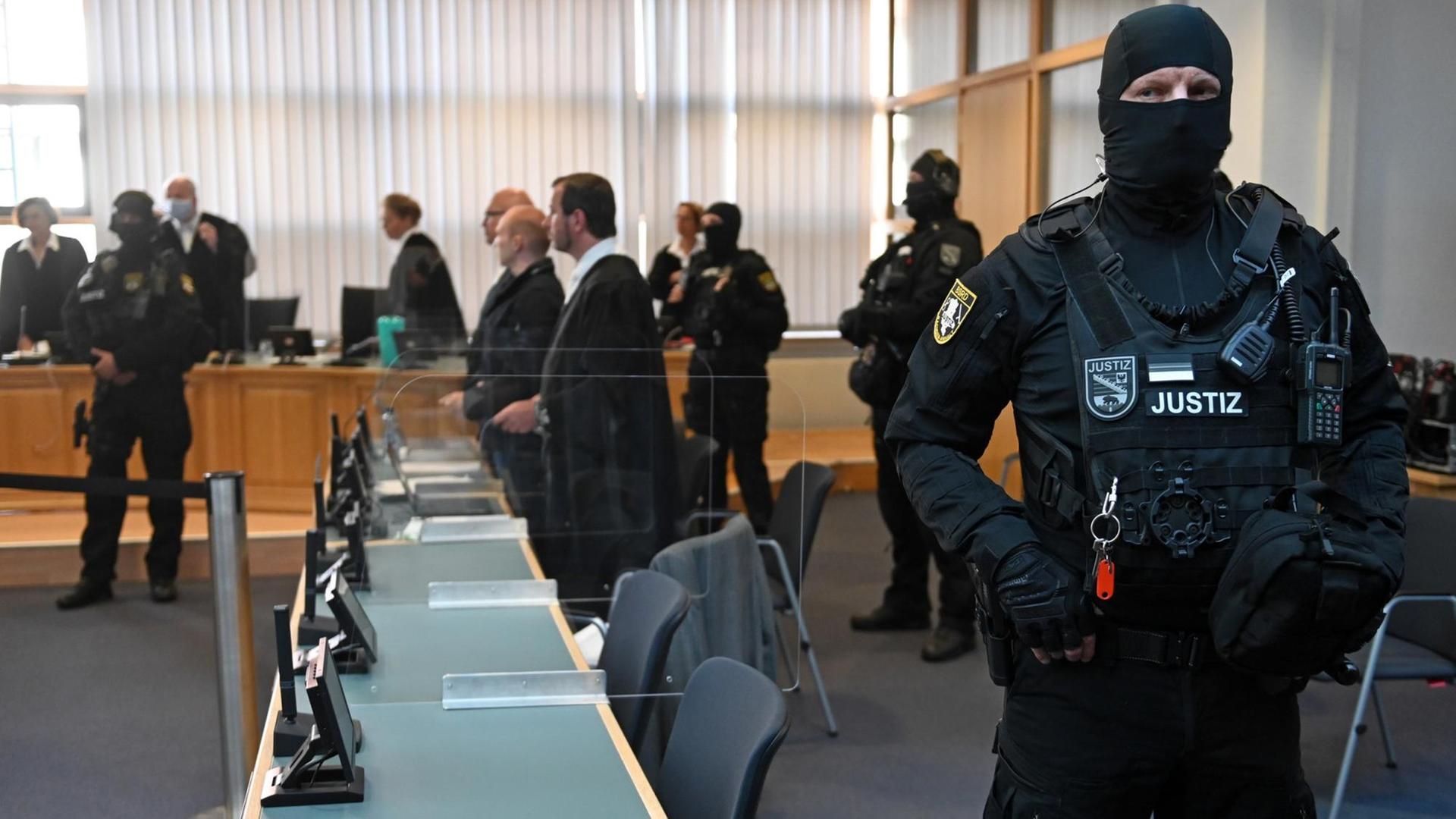 In einem Gerichtsaal steht im Vordergrund ein bewaffneter Justizbeamter. Im Hintergrund sind zu seheh: der Angeklagte, der Verteidiger, das Gericht und weitere vermummte und bewaffnete Justizbeamte.