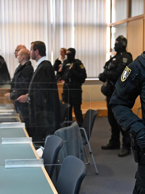 In einem Gerichtsaal steht im Vordergrund ein bewaffneter Justizbeamter. Im Hintergrund sind zu seheh: der Angeklagte, der Verteidiger, das Gericht und weitere vermummte und bewaffnete Justizbeamte.