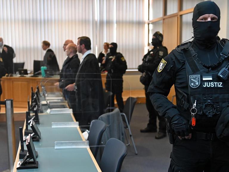 In einem Gerichtsaal steht im Vordergrund ein bewaffneter Justizbeamter. Im Hintergrund sind zu seheh: der Angeklagte, der Verteidiger, das Gericht und weitere vermummte und bewaffnete Justizbeamte.