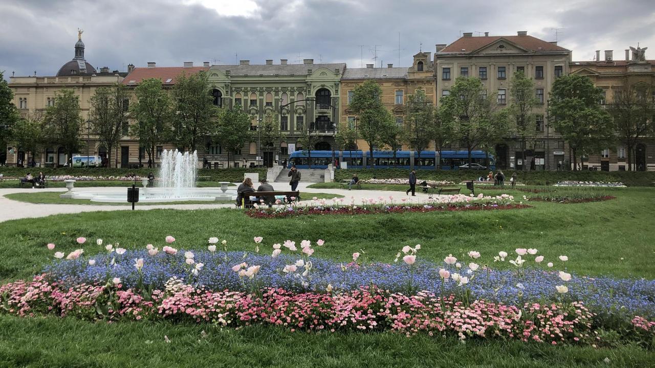 Blick auf einen Park. Im Vordergrund ein großes Beet mit blauen, weißen und rosa blühenden Blumen, dahinter ein Springbrunnen. Im Hintergrund Häuserfassaden.