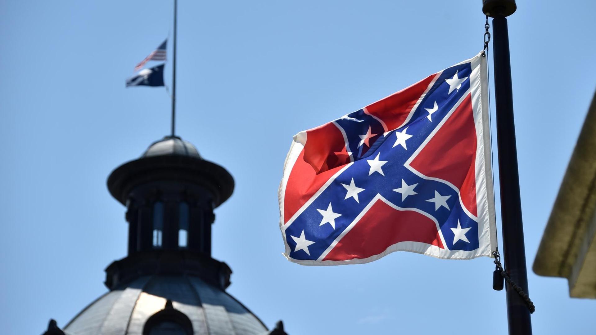 Die Südstaaten-Flagge weht vor blauem Himmel; im Hintergrund sieht man die Kuppel eines Gebäudes.