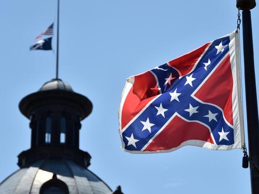 Die Südstaaten-Flagge weht vor blauem Himmel; im Hintergrund sieht man die Kuppel eines Gebäudes.