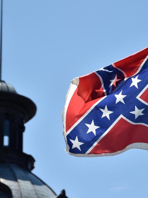 Die Südstaaten-Flagge weht vor blauem Himmel; im Hintergrund sieht man die Kuppel eines Gebäudes.