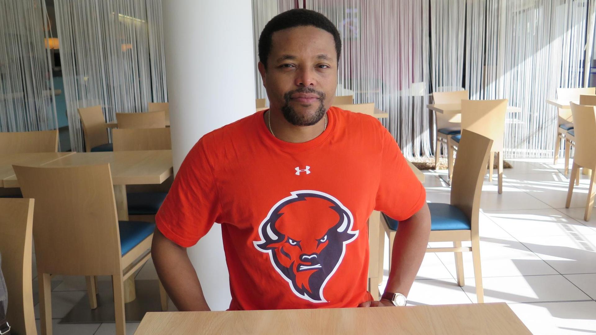 Ein Mann im orangen T-Shirt sitzt in einem leeren Restaurant und schaut lächelnd in die Kamera. Ein Mann im orangen T-Shirt sitzt in einem leeren Restaurant und schaut lächelnd in die Kamera.
