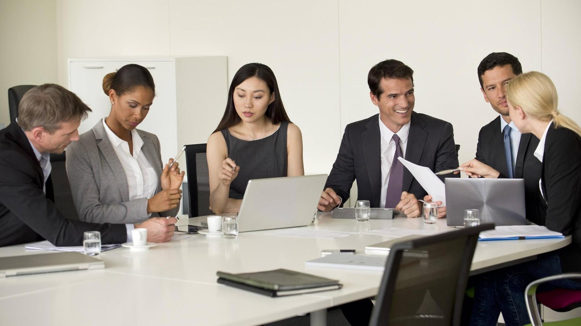 Drei Männer und drei Frauen in Business-Kleidung sitzen um einen Konferenz-Tisch, auf dem Laptops stehen Drei Männer und drei Frauen in Business-Kleidung sitzen um einen Konferenz-Tisch, auf dem Laptops stehen