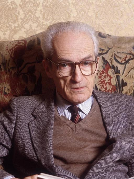 Ein alter Mann mit weißen Haaren und mit Schlips und großer Brille sitzt auf einem altmodisch wirkenden Sofa und hält ein geöffnetes Buch im Schoß.