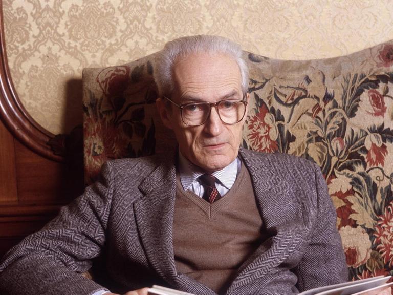 Ein alter Mann mit weißen Haaren und mit Schlips und großer Brille sitzt auf einem altmodisch wirkenden Sofa und hält ein geöffnetes Buch im Schoß.