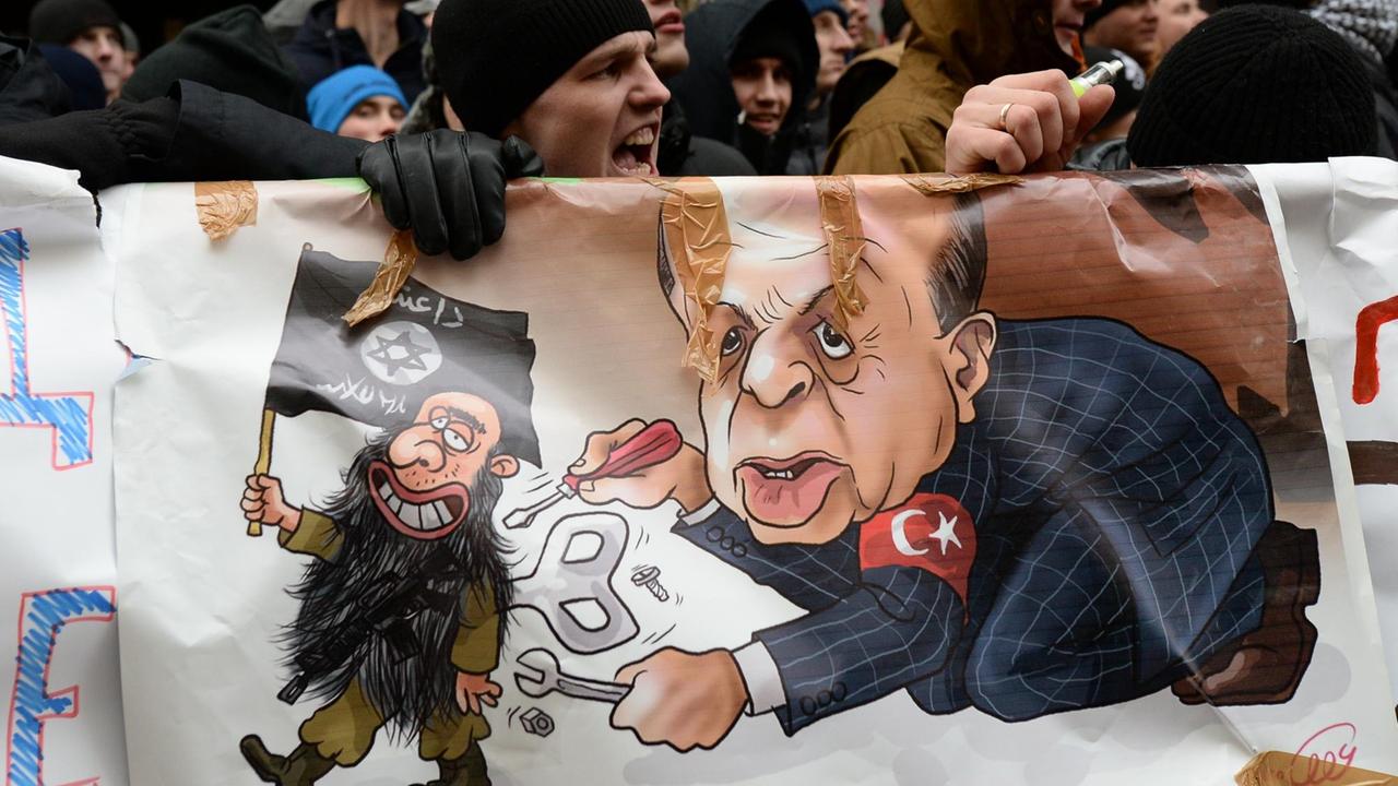 Demonstranten halten ein Plakat mit einer Zeichnung des türkischen Präsidenten Erdogan, der ein Terroristen-Männchen mit IS-Fahne "aufzieht". Demonstranten halten ein Plakat mit einer Zeichnung des türkischen Präsidenten Erdogan, der ein Terroristen-Männchen mit IS-Fahne "aufzieht".