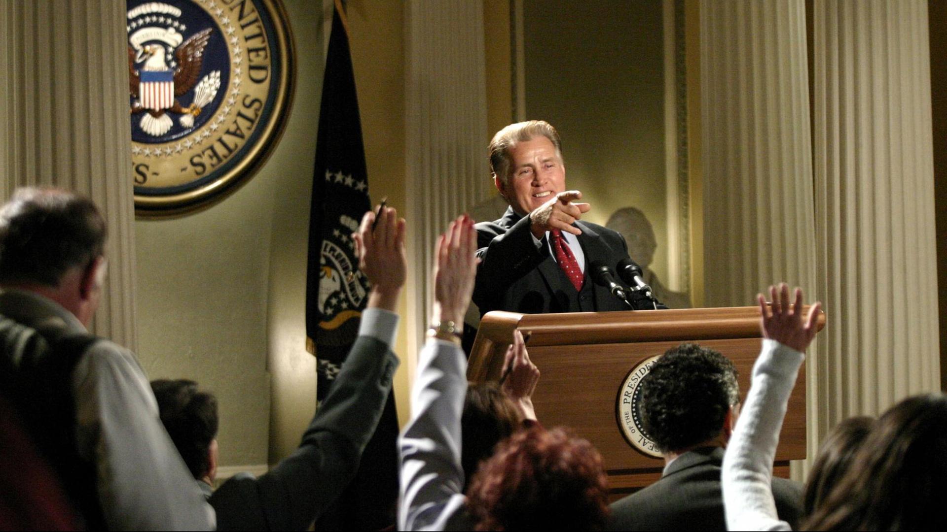 Martin Sheen steht als Präsident Josiah Bartlet in 'The West Wing' vor einem Rednerpult und zeigt auf jemanden in der Menge. Martin Sheen steht als Präsident Josiah Bartlet in 'The West Wing' vor einem Rednerpult und zeigt auf jemanden in der Menge.