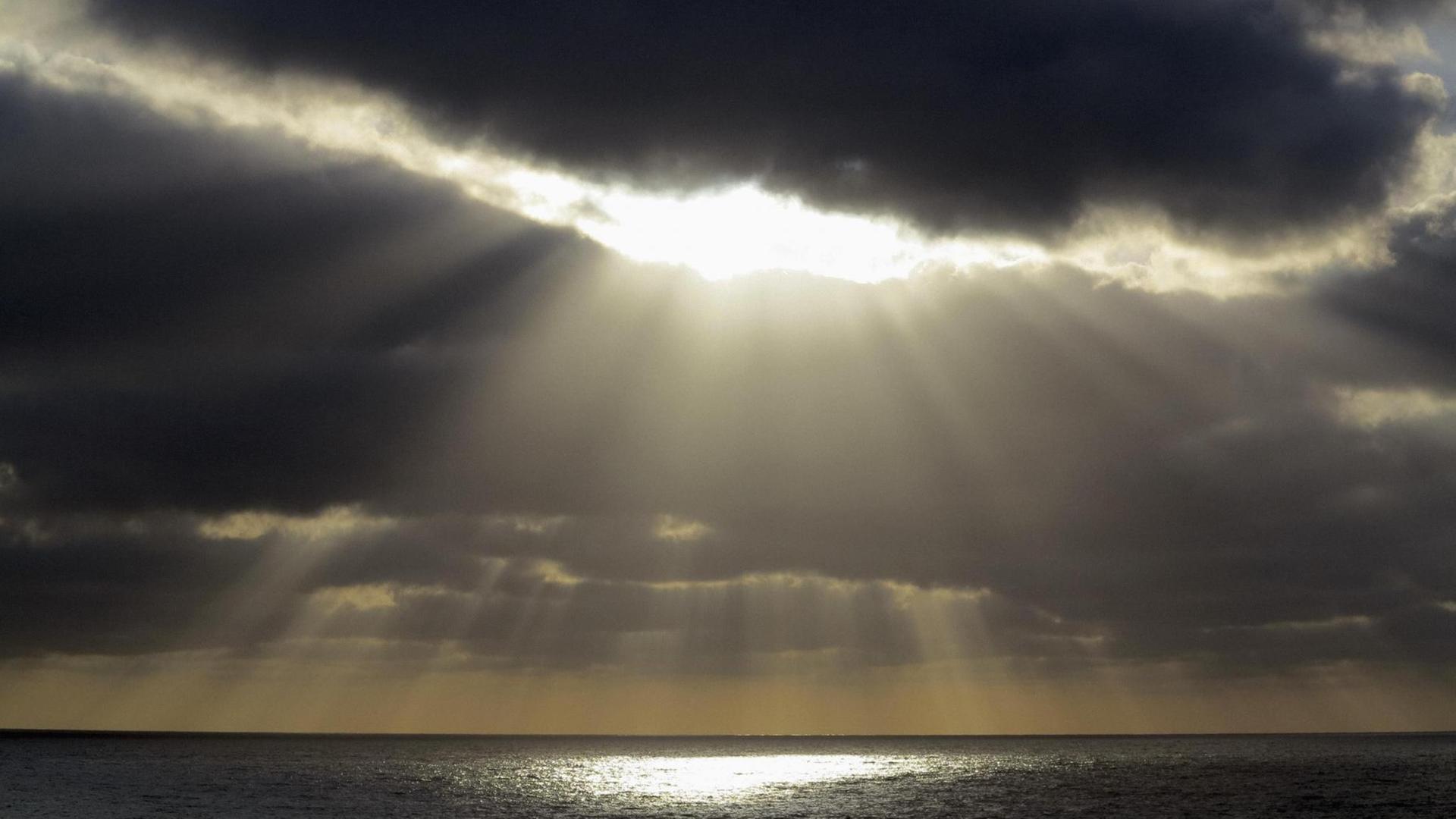 Sonnenstrahlen brechen durch eine dunkle Wolke über dem Ozean und spiegeln sich darin. Sonnenstrahlen brechen durch eine dunkle Wolke über dem Ozean und spiegeln sich darin.