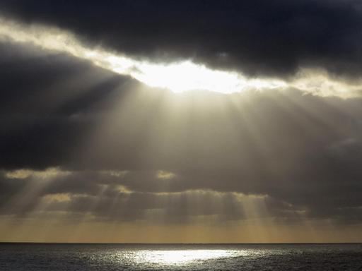 Sonnenstrahlen brechen durch eine dunkle Wolke über dem Ozean und spiegeln sich darin.