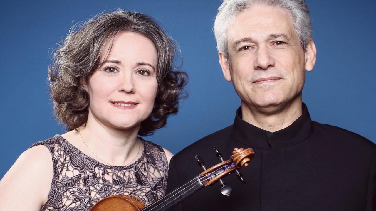 Judith Ingolfsson und Vladimir Stoupel stehen vor einem blauen Hintergrund und schauen in die Kamera. Judith Ingolfsson hält dabei ihre Geige in ihren Händen. Judith Ingolfsson und Vladimir Stoupel stehen vor einem blauen Hintergrund und schauen in die Kamera. Judith Ingolfsson hält dabei ihre Geige in ihren Händen.