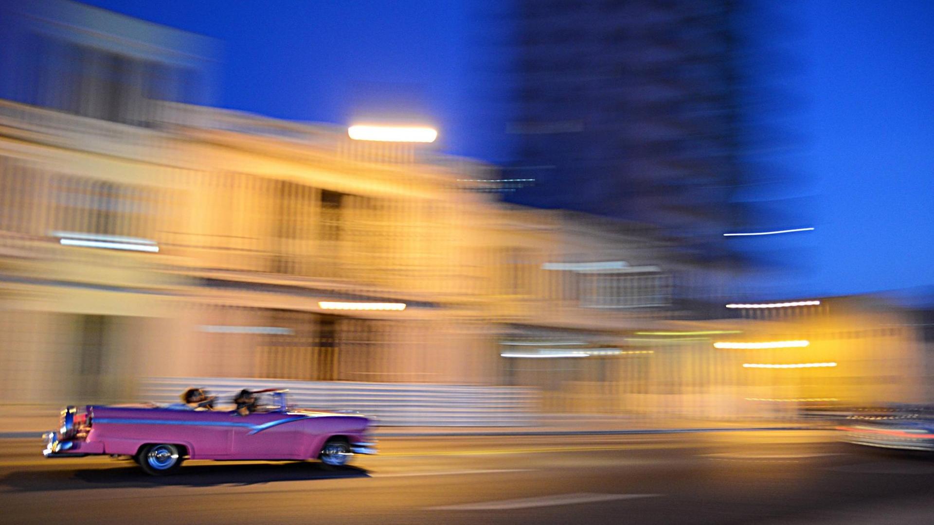 Ein pinker Oldtimer rauscht im Abendlicht vor verwischtem Hintergrund an einer hellen Häuserzeile vorbei.