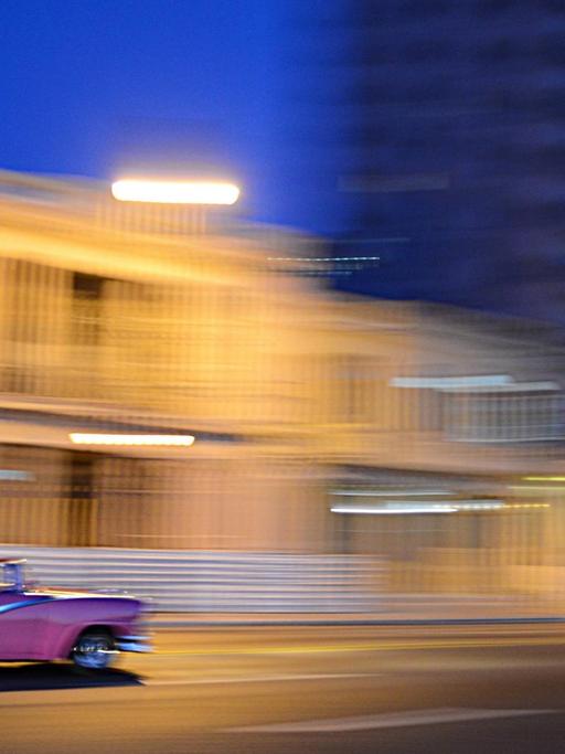 Ein pinker Oldtimer rauscht im Abendlicht vor verwischtem Hintergrund an einer hellen Häuserzeile vorbei.