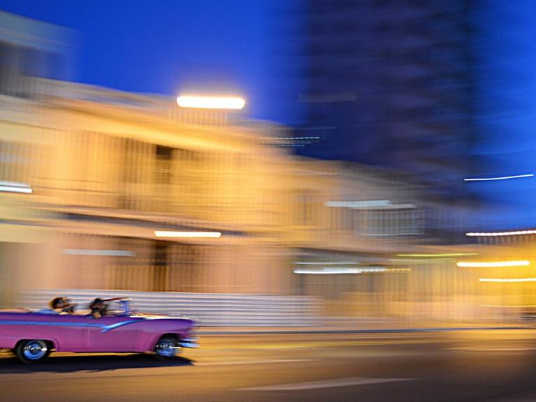 Ein pinker Oldtimer rauscht im Abendlicht vor verwischtem Hintergrund an einer hellen Häuserzeile vorbei.