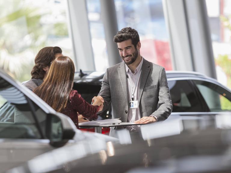 Ein Autoverkäufer schüttelt die Hände seiner Kunden - mit einem Lächeln im Gesicht.