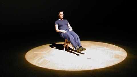 Eine Performerin sitzt im hellen Lichtkegel einer dunklen Bühne auf einem Stuhl und streckt die Füße