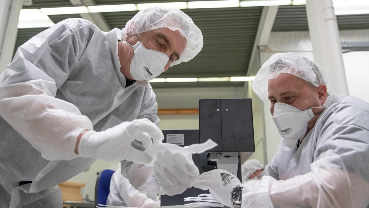 Zwei Männer in weißer Schutzkleidung hantieren in einem Werkraum mit Schutzmasken. Zwei Männer in weißer Schutzkleidung hantieren in einem Werkraum mit Schutzmasken.