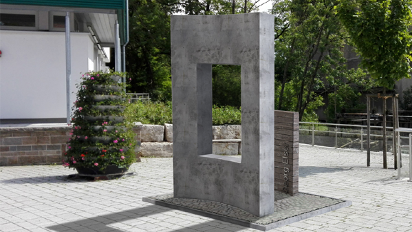 Das Bild zeigt das Denkmal für Georg Elser, eine aufrecht stehende Betonscheibe mit einem Ausschnitt. Daneben eine Holzscheibe, die den Ausschnitt füllen würde.