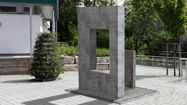 Das Bild zeigt das Denkmal für Georg Elser, eine aufrecht stehende Betonscheibe mit einem Ausschnitt. Daneben eine Holzscheibe, die den Ausschnitt füllen würde. Das Bild zeigt das Denkmal für Georg Elser, eine aufrecht stehende Betonscheibe mit einem Ausschnitt. Daneben eine Holzscheibe, die den Ausschnitt füllen würde.