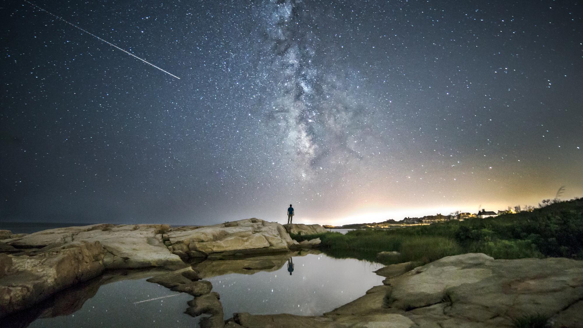 Ein Mensch steht vor einem Sternenhimmel, auf dem eine Sternschnuppe zu sehen ist.