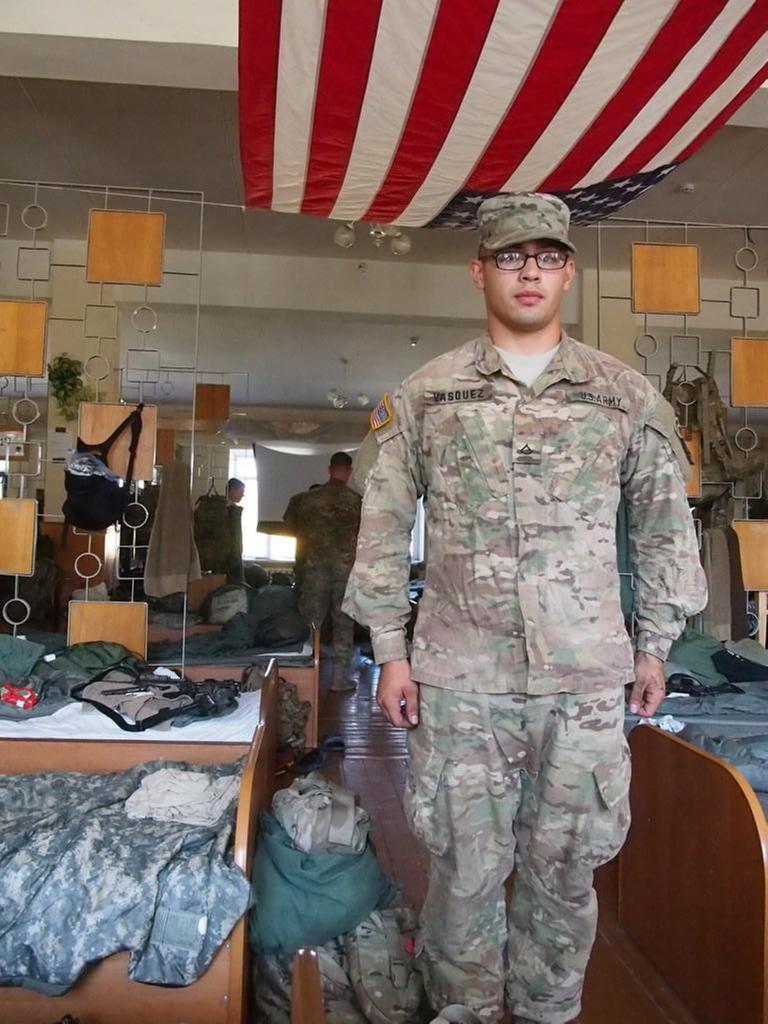 US-amerikanischer Soldat steht in einem Schlafsaal stramm, aber der Decke eine US-Flagge, im Hintergrund Betten.