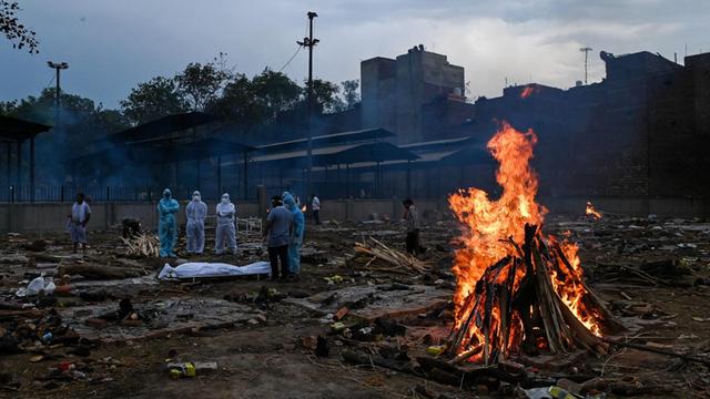 Menschen in Schutzanzügen stehen in der Nähe eines toten Angehörigen, der am Coronavirus gestorben ist, auf einem Einäscherungsgelände in Neu-Delhi. Im rechten Bildvordergrund ist ein Feuer zu sehen.