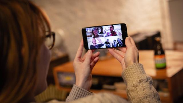 Eine Frau hat ein Smartphone der Hand, auf dem Bildschirm sind sechs Verwandte oder Freunde zu sehen, mit denen sie per Video kommuniziert.