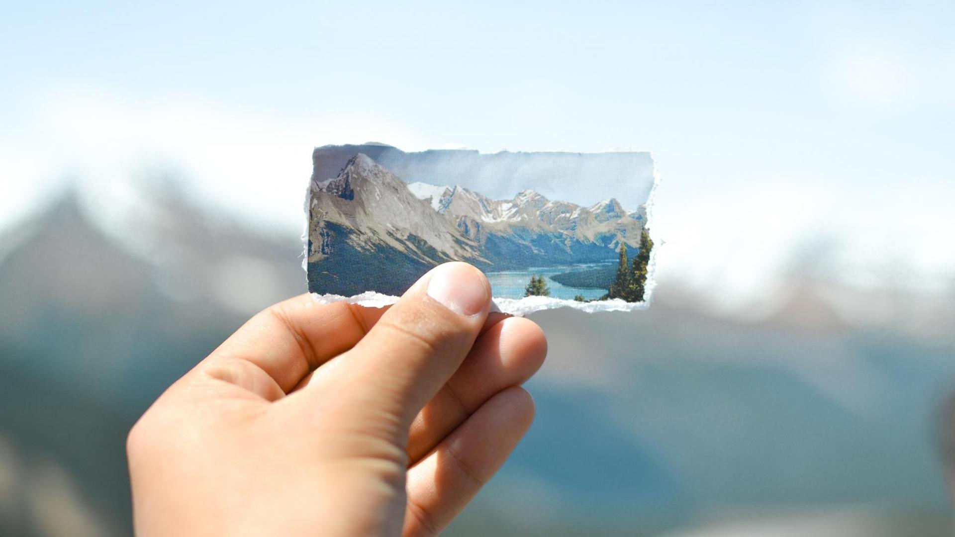 Eine Hand hält ein ausgerissenes Bild von Eisgletschern und Bergen vor die Silhouette der echten Berge.
