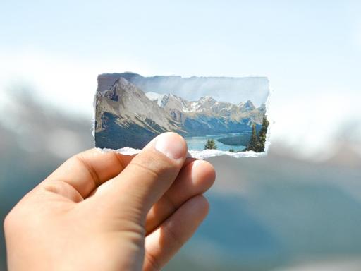 Eine Hand hält ein ausgerissenes Bild von Eisgletschern und Bergen vor die Silhouette der echten Berge.
