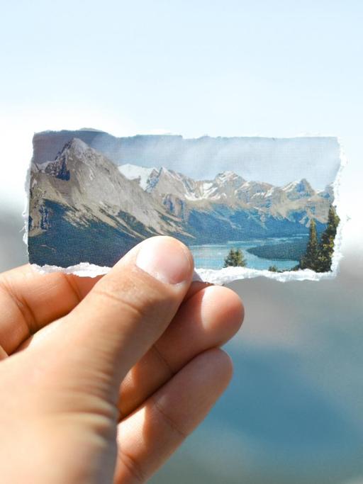 Eine Hand hält ein ausgerissenes Bild von Eisgletschern und Bergen vor die Silhouette der echten Berge.