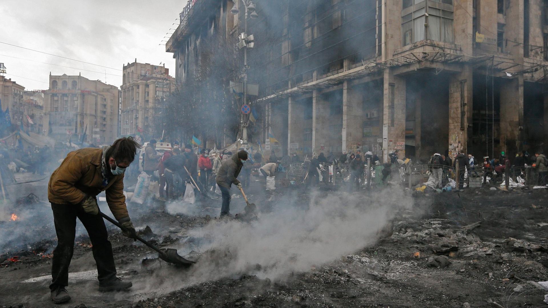 Auf dem verwüsteten Unabhängigkeitsplatz in Kiew versuchen mehrere Männer mit Schaufeln, qualmenden Schutt zu entfernen.