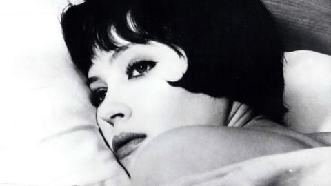 Eine Filmszene aus "Die Geschichte der Nana S." von Jean Luc Godard zeigt Anna Karina in einer Nahaufnahme auf einem weißen Kissen liegend.