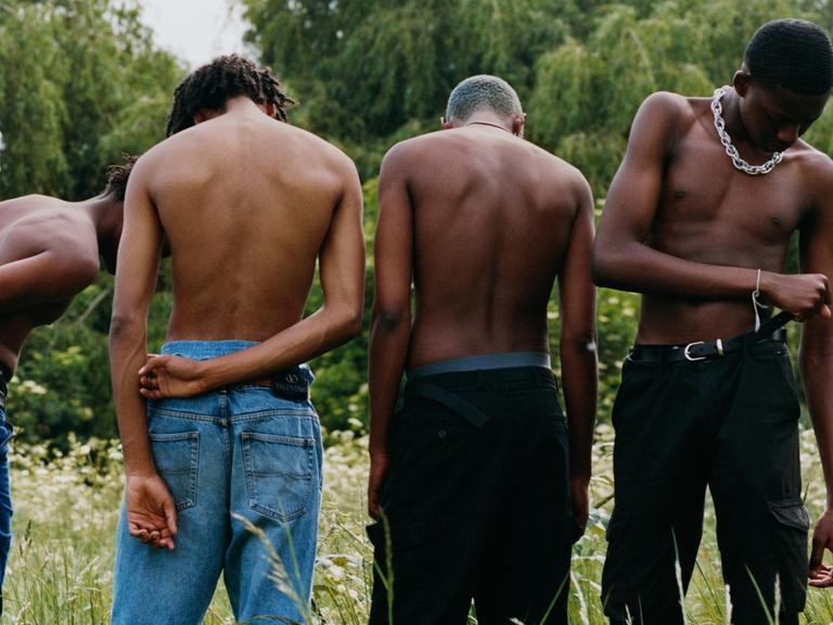 Fünf schwarze Jungs stehen oberkörperfrei in einer Wiese.