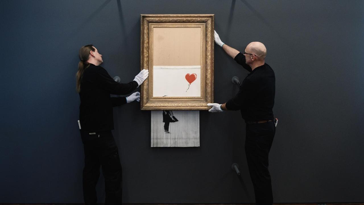 Zwei in schwarz gekleidete Museumsmitarbeiter hängen mit weißen Handschuhen Banksys Bild "Love in the Bin" an die Wand. Die Hälfte des Bildes hängt halb geschreddert aus dem goldenen Rahmen heraus. Zwei in schwarz gekleidete Museumsmitarbeiter hängen mit weißen Handschuhen Banksys Bild "Love in the Bin" an die Wand. Die Hälfte des Bildes hängt halb geschreddert aus dem goldenen Rahmen heraus.