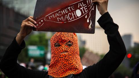 Eine Demonstrantin, in einer Leopardenmuster-Maske, bei Protesten gegen Femizide in Mexiko Stadt, mit einem Schild mit der Aufschrift "Machete all Machote!!!"